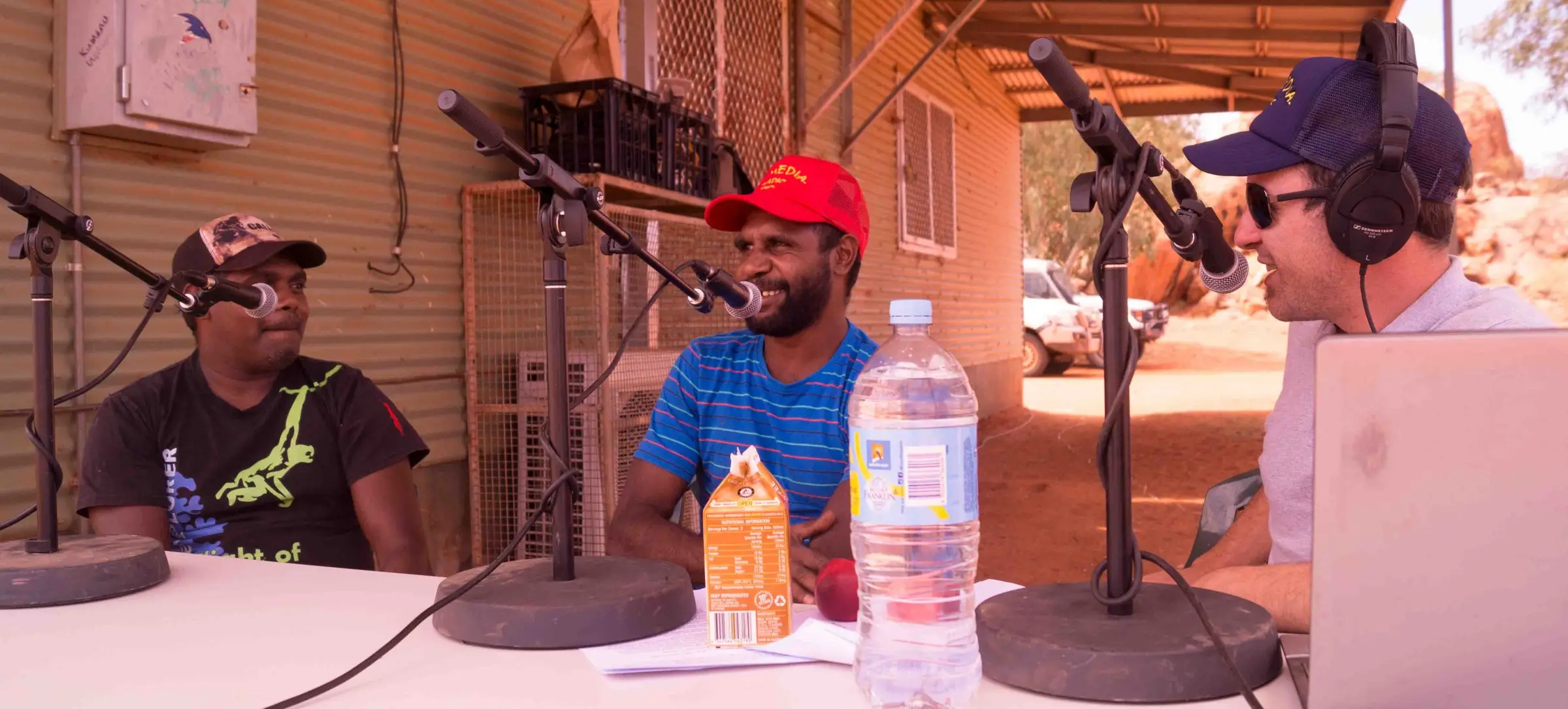 Three people at an outside broadcast table on red earth