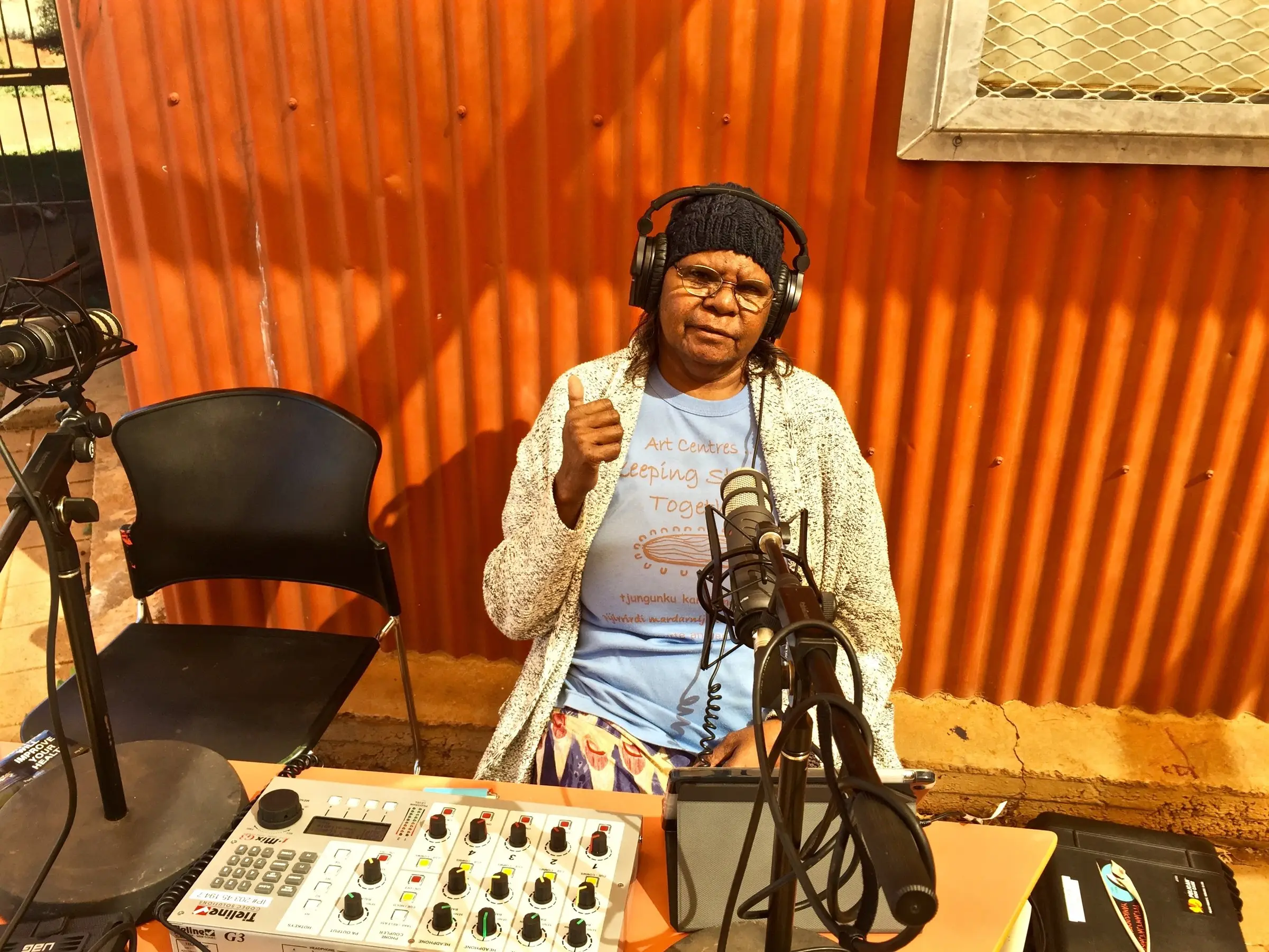 A Radio 5NPY broadcaster with headphones at the microphone, in front of the orange studio wall at Umuwa
